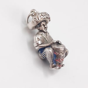 May include: A silver charm depicting a musician playing a drum. The charm features a figure wearing a hat with the word "Nassau" on it, a shirt, and striped pants. The drum has red and blue accents.