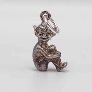 May include: A silver charm in the shape of a seated goblin or elf, with a loop for attaching to a bracelet or necklace. The figure is detailed with facial features and is hugging its knees.