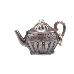Puede incluir: Un pequeño dije de tetera plateada con detalles intrincados. La tetera presenta un pico curvo, un cuerpo redondeado con líneas verticales y un lazo para sujetar. La tapa tiene un patrón decorativo.