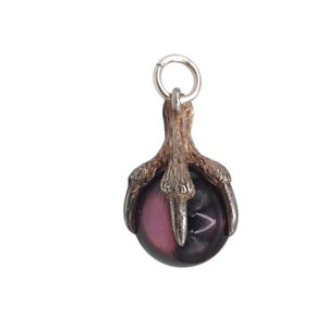 May include: A pendant featuring a silver-toned claw grasping a round, two-toned gemstone. The gemstone is a mix of dark purple and pink, with a silver-toned loop at the top for hanging. The pendant is set against a white background.