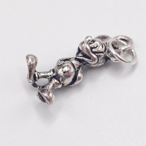 Pode incluir: Um berloque de metal em tom prateado apresentando uma figura tridimensional de um rato de desenho animado com orelhas grandes, vestindo shorts e sapatos, com um pequeno anel circular preso ao topo da cabe&ccedil;a para pendurar.