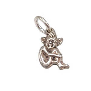 May include: A silver-colored charm in the shape of a seated goblin or elf. The charm has a loop at the top for attachment and is detailed with facial features and a pointed hat. The goblin is hugging its knees.