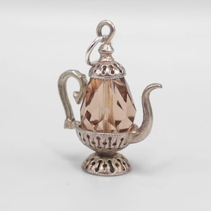 Puede incluir: Un charm de tetera plateado con un cuerpo de cristal facetado de color melocotón. El charm presenta un asa y un pico curvos, y una base decorativa. Es una joya.