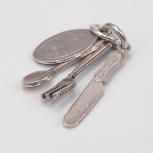 Op de afbeelding: Een zilveren bedel met miniatuur bestek: een lepel, vork en mes, samen met een kleine muntvormige hanger. De bedel is bevestigd aan een zilveren ring. De items zijn gedetailleerd en lijken van sterling zilver te zijn.