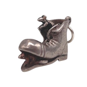 May include: A silver-toned charm in the shape of a boot with a mouse peeking out. The boot has a rounded toe and a loop for attaching to a keychain or necklace. The mouse is detailed with small ears and a tail.