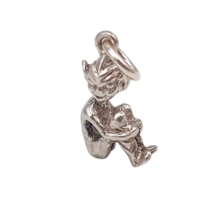 May include: A silver-toned charm depicting a seated pixie figure. The pixie is curled up, with its knees drawn up and arms wrapped around them. The charm has a loop at the top for attachment to a necklace or bracelet.