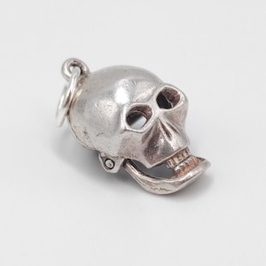 May include: A silver skull charm with a hinged jaw and a loop for attaching to a necklace or bracelet. The skull has detailed features, including eye sockets, a nose cavity, and teeth. The charm is likely made of metal and is designed to be a pendant.