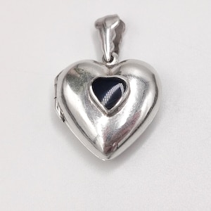 May include: A silver heart-shaped locket pendant with a small, dark blue stone in the centre. The locket has a hinged design and a loop for a chain. The pendant is on a white background.