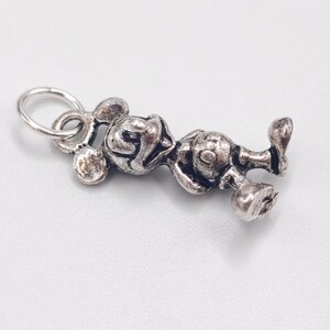 Pode incluir: Um pingente de metal prateado em forma de personagem de rato de desenho animado com orelhas grandes e redondas, rosto sorridente e corpo arredondado. O pingente apresenta um acabamento texturizado e envelhecido com um pequeno anel circular.