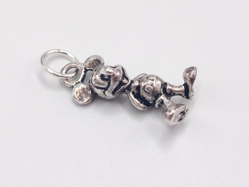 Pode incluir: Um pingente de metal prateado em forma de personagem de rato de desenho animado com orelhas grandes e redondas, rosto sorridente e corpo arredondado. O pingente apresenta um acabamento texturizado e envelhecido com um pequeno anel circular.