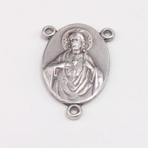 Puede incluir: Colgante religioso plateado de forma ovalada. El colgante presenta un relieve detallado de una figura con halo, manos en el pecho y un corazón. El colgante tiene tres pequeños lazos para la fijación.
