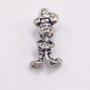 Pode incluir: Um berloque de metal em tom prateado com o formato de uma figura de rato de desenho animado em p&eacute;. A parte de tr&aacute;s da cabe&ccedil;a apresenta o texto em relevo DISNEY e 925. O berloque inclui um pequeno anel circular no topo para fixa&ccedil;&atilde;o.
