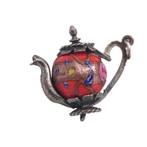 Puede incluir: Broche decorativo con forma de tetera. La tetera tiene un cuerpo rojo, como de cristal, con diseños florales y abstractos de colores. El asa, el pico, la tapa y la base son de metal plateado. Un accesorio único.