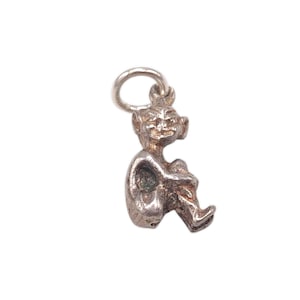 May include: A small, silver-toned charm in the shape of a seated goblin or elf. The charm has a loop at the top for attachment and features detailed facial and body features. The figure is in a seated position with its knees drawn up.