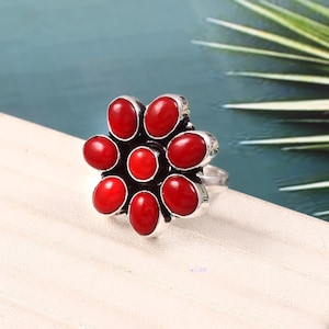 Puede incluir: Un anillo de plata con un diseño en forma de flor, engastado con ocho piedras ovaladas de color rojo brillante. La banda del anillo es plateada y las piedras están dispuestas en un patrón floral. El anillo se muestra sobre una superficie blanca.
