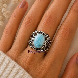 May include: A silver ring featuring an oval, light blue gemstone. The ring has an ornate, filigree design around the stone and the text "FizaJewels" is visible on the ring.