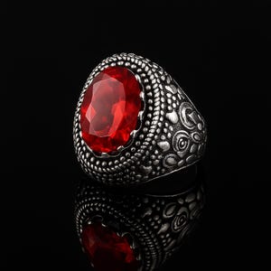 May include: A silver-toned ring with an oval, faceted, red gemstone. The ring's band features a detailed, textured design with small circular and floral patterns. The ring is displayed on a reflective black surface, highlighting its intricate details.