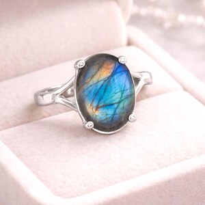 Puede incluir: Anillo de plata con una piedra labradorita ovalada. La piedra muestra un vibrante juego de colores, con tonos azules, naranjas y amarillos. La banda del anillo tiene un diseño dividido, lo que aumenta su elegancia. El anillo está en una caja de joyería.