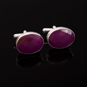 May include: A pair of silver cufflinks featuring oval-cut, deep magenta gemstones. The cufflinks are set against a black background, with reflections visible below the jewellery.