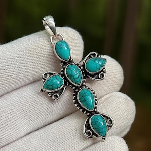 May include: A silver cross pendant with five turquoise gemstones. The cross design includes decorative swirls around each stone. The turquoise stones are teardrop-shaped and have a natural, mottled pattern. The pendant has a loop for a chain.