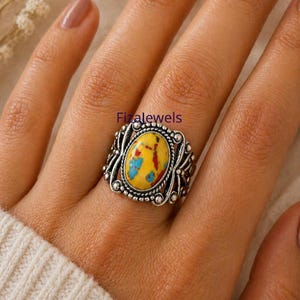 May include: A silver ring with an oval yellow stone, speckled with blue and red. The stone is set in an ornate frame. The ring is on a hand, with the brand name "Fizalewels" visible. The ring's design is intricate.