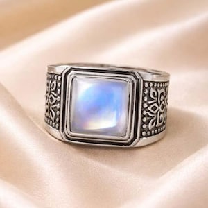 Könnte beinhalten: Silberfarbener Ring mit einem quadratischen, irisierenden, hellblauen Edelstein in der Mitte. Der Ring hat ein verziertes, graviertes Design um den Edelstein und auf dem Band, was eine Vintage- oder Antik-Ästhetik erzeugt.