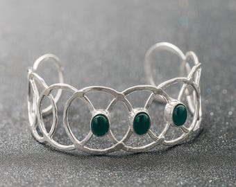 May include: A silver cuff bracelet with a geometric design featuring three oval, dark green gemstones. The bracelet has an open design with interconnected circles and a hammered texture, set against a dark, speckled background.