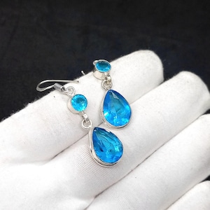Peut inclure: Boucles d'oreilles en argent ornées de pierres précieuses bleues vives. Chaque boucle d'oreille est dotée d'un crochet et présente une pierre ronde et une pierre plus grande en forme de goutte. Les pierres sont facettées.