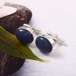 May include: Silver cufflinks featuring oval lapis lazuli stones in a deep blue hue, speckled with gold. The stones are set in a polished silver frame. The cufflinks are presented on a light surface, with a green leaf and a dark stone visible in the background.