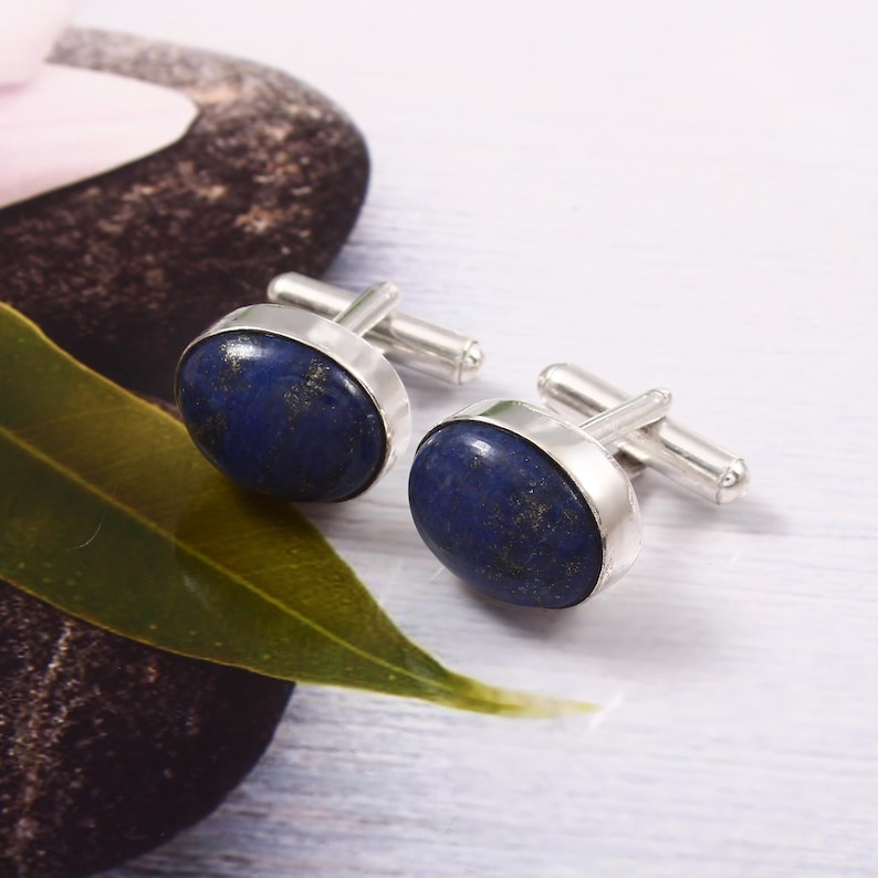 May include: Silver cufflinks featuring oval lapis lazuli stones in a deep blue hue, speckled with gold. The stones are set in a polished silver frame. The cufflinks are presented on a light surface, with a green leaf and a dark stone visible in the background.