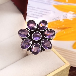 Puede incluir: Un anillo de plata con un diseño floral con ocho piedras de amatista ovaladas de color púrpura intenso. El anillo está engastado en una banda de plata y se muestra en una caja de joyería.
