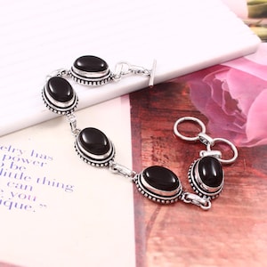 May include: Silver-toned bracelet featuring oval black gemstones. The bracelet has a toggle clasp and is set on a light-colored surface. The gemstones are framed with a decorative border.