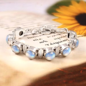 May include: A silver bracelet with oval, iridescent moonstone cabochons. The bracelet has a toggle clasp and is set on a vintage paper background. The moonstones have a soft, milky blue and white colour.