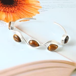 May include: A silver-toned cuff bracelet with five teardrop-shaped tiger's eye stones. The bracelet has a wavy design and is set against a soft, light background with an orange flower in the upper left corner.