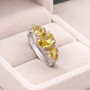 Puede incluir: Un anillo plateado con tres gemas amarillas. La piedra central, de forma ovalada, está flanqueada por dos piedras en forma de pera. El anillo se destaca sobre un fondo suave y neutro, resaltando el color vibrante de las gemas.