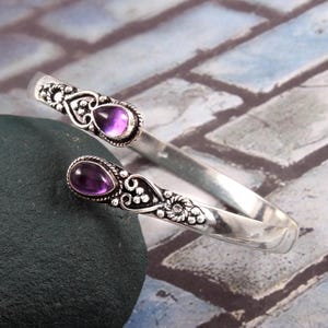 Peut inclure: Bracelet manchette argenté orné de deux pierres d'améthyste violettes ovales. Le bracelet ouvert présente des détails floraux et en forme de cœur. Les pierres sont serties dans un sertissage décoratif.