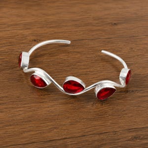 Puede incluir: Una pulsera tipo brazalete de plata con un diseño de banda ondulada y abierta. La pulsera está decorada con cuatro piedras facetadas de color rojo intenso en forma de lágrima, engastadas a lo largo del marco plateado ondulante.