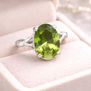 May include: A silver ring with a large, oval-cut peridot gemstone. The peridot is a vibrant, light green colour and is set in a four-prong setting. The ring is displayed in a light pink jewellery box.