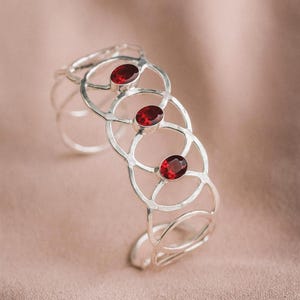 Op de afbeelding: Een zilveren manchetarmband met een open ontwerp van onderling verbonden cirkels. Elke cirkel bevat een levendige, ovaalvormige, rode edelsteen. De armband heeft een gepolijste afwerking en een klassieke, elegante stijl.