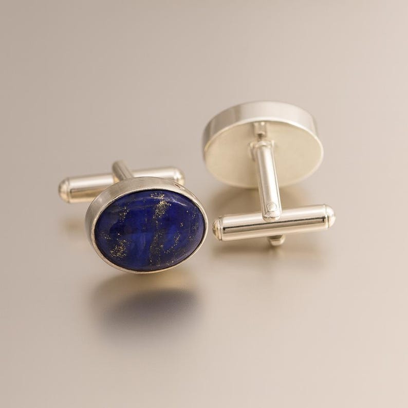 May include: Silver cufflinks featuring oval, deep blue lapis lazuli stones with gold inclusions. The cufflinks have a T-bar fastening. The background is a light grey colour.