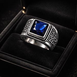 May include: A silver-toned ring with a large, square, deep blue gemstone. The ring has an ornate, engraved design on the band and is presented in a black ring box. The gemstone is the focal point of the ring.