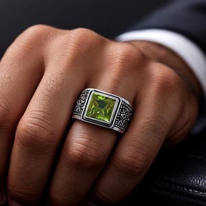 May include: A silver ring with a square, light green gemstone. The ring has an ornate design on the band. The ring is shown on a person's hand.
