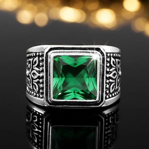 May include: A silver-toned ring with a large, square, emerald-green gemstone at the center. The band features ornate, symmetrical patterns on the sides. The ring is set against a dark, reflective surface, highlighting the gemstone's brilliance.