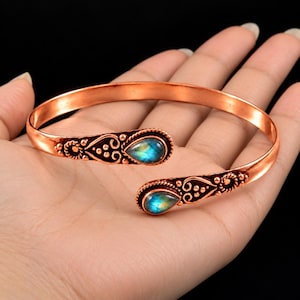 May include: A copper cuff bracelet featuring an open design with two teardrop-shaped labradorite gemstones at the ends. The metal surface is decorated with intricate embossed scrollwork and small beaded details surrounding each stone.