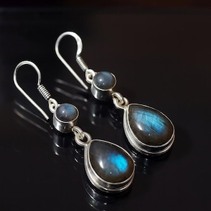 Op de afbeelding: Zilveren oorhangers met labradoriet edelstenen. Elke oorbel heeft een kleine, ronde steen en een grotere, druppelvormige steen met blauwgroene iriserende glans. De oorbellen hebben een haaksluiting.