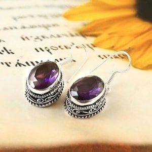 Può includere: Un paio di orecchini color argento con pietre preziose ovali viola scuro. Gli orecchini hanno un bordo decorativo a motivi e una chiusura a gancio. Lo sfondo è di un colore tenue e neutro, con un accenno di fiore giallo.
