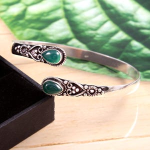 Op de afbeelding: Zilverkleurige manchetarmband met groene, druppelvormige edelstenen. De armband heeft sierlijke details rond de edelstenen en langs de uiteinden, met een bloemen- en stippenpatroon. De armband is open en verstelbaar.
