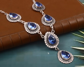 May include: A silver-toned necklace featuring teardrop-shaped blue gemstones. The gemstones are set within a detailed, circular frame. The necklace is displayed on a brown surface with a teal background, showcasing the jewelry's design and color.