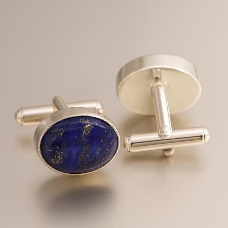 May include: Silver cufflinks featuring oval, deep blue lapis lazuli stones with gold flecks. The cufflinks have a classic design with a post and toggle closure.
