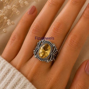 May include: A silver ring featuring an oval, yellow gemstone. The ring has an intricate, filigree design and the text "Fizajewels". The ring is on a person's finger.